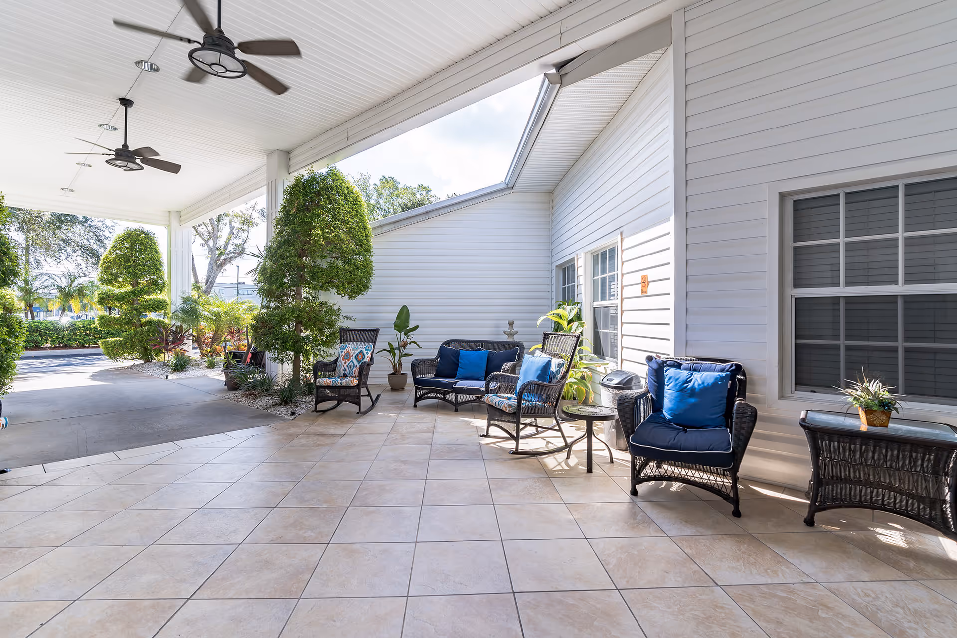 Covered outdoor patio area with tiled floor, ceiling fans, and wicker seating including chairs and a loveseat with blue cushions. There are potted plants and neatly trimmed bushes along the edge, with white siding walls and windows in the background.