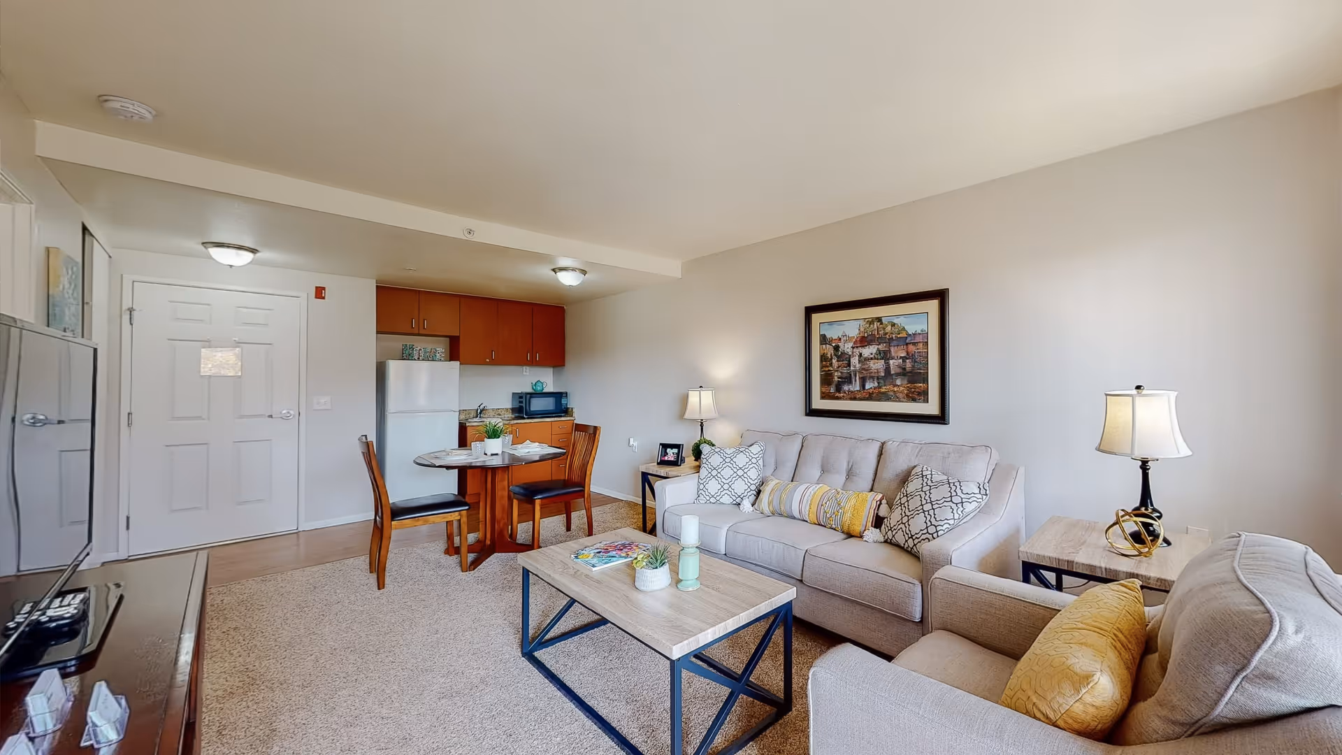 A cozy living room and dining area in a senior living facility named Citrus Place. The room features a beige sofa with patterned and yellow pillows, a matching armchair, a wooden coffee table with decorative items, and a side table with a lamp. There is a round dining table with two chairs near a small kitchen area equipped with a refrigerator, microwave, and cabinets. The walls are light-colored with a framed painting above the sofa, and the floor is carpeted.