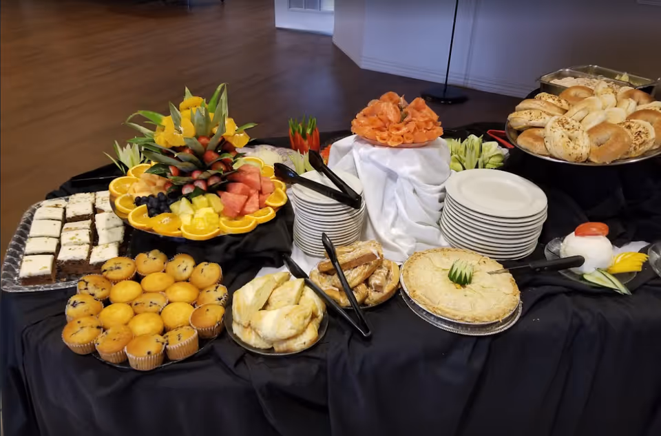 Buffet table with plates, bagels, muffins, pastries, and a fruit platter arranged on a black tablecloth in an indoor dining area.