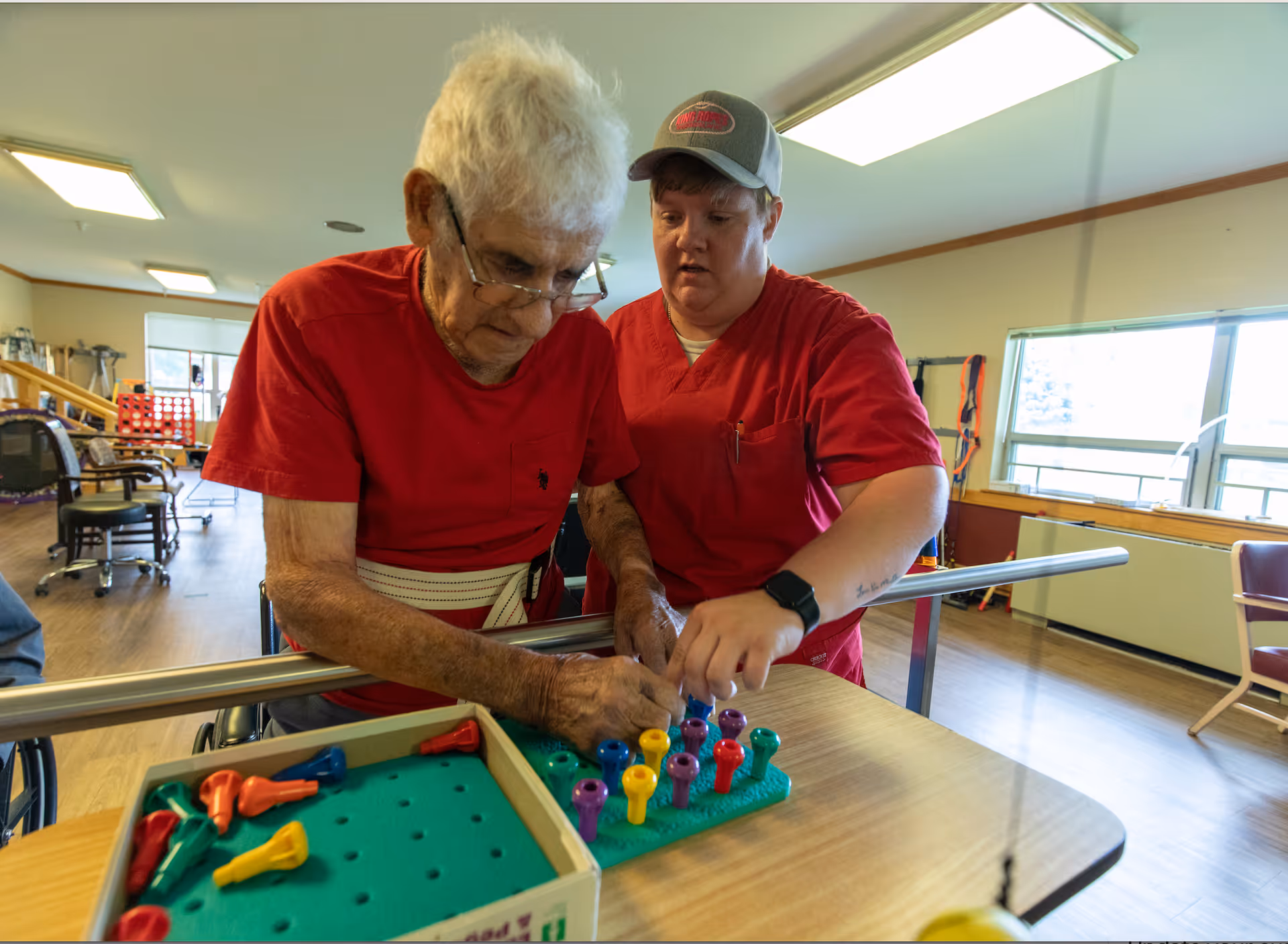 An elderly man wearing a red shirt and glasses is engaged in a pegboard activity with the assistance of a caregiver in a red uniform and cap inside a well-lit room with wooden flooring and large windows.
