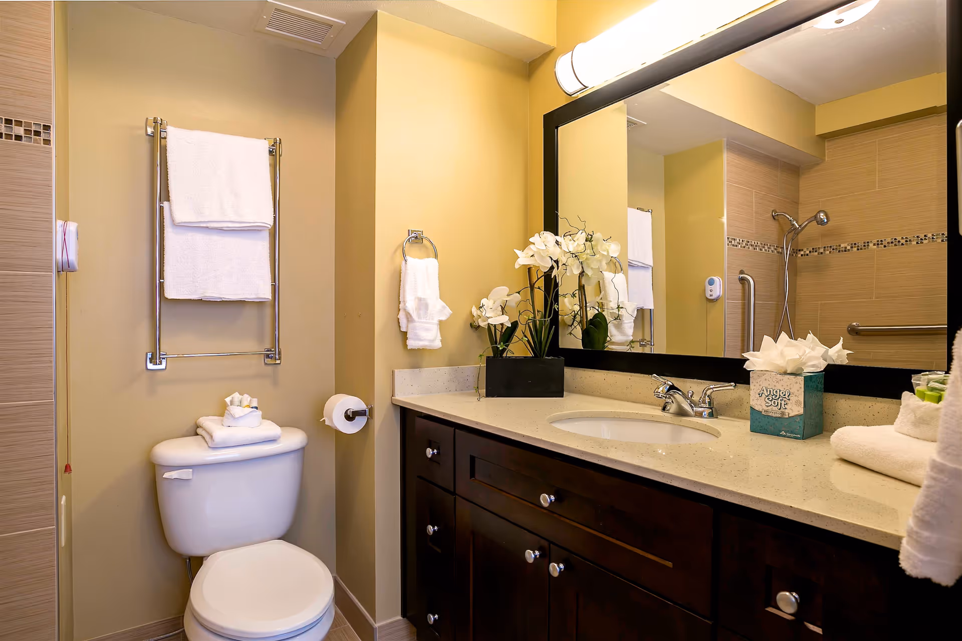 A clean and well-lit bathroom featuring a white toilet with folded towels on top, a towel rack with white towels above the toilet, a beige wall, a dark wood vanity with a white countertop and an oval sink, a large mirror with a light fixture above it, a box of Angel Soft tissues, and a decorative arrangement of white flowers. The shower area with a handheld showerhead and grab bars is visible in the mirror reflection.