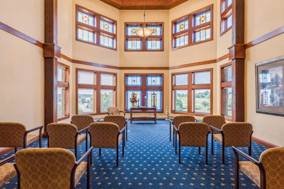A bright room with high ceilings and multiple stained glass windows. The room has blue carpet with a patterned design and several upholstered chairs arranged in rows facing a small wooden table with floral arrangements and candles. The walls are cream-colored with wooden trim and beams, and there is a hanging light fixture from the ceiling.