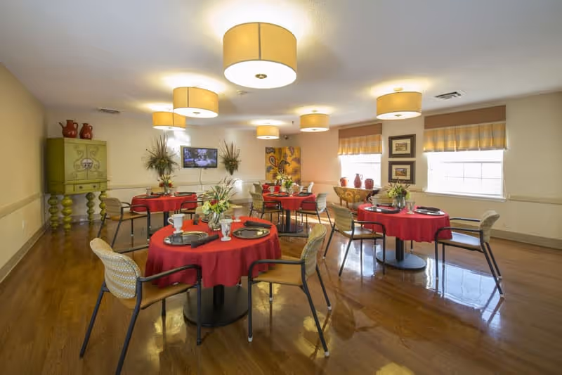 Bright dining room with round tables set with red tablecloths and chairs, overhead drum lights, decorative cabinets and windows.