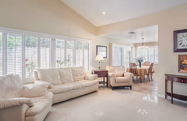 Bright living room with white sofas, large windows with shutters, and a dining area in back.