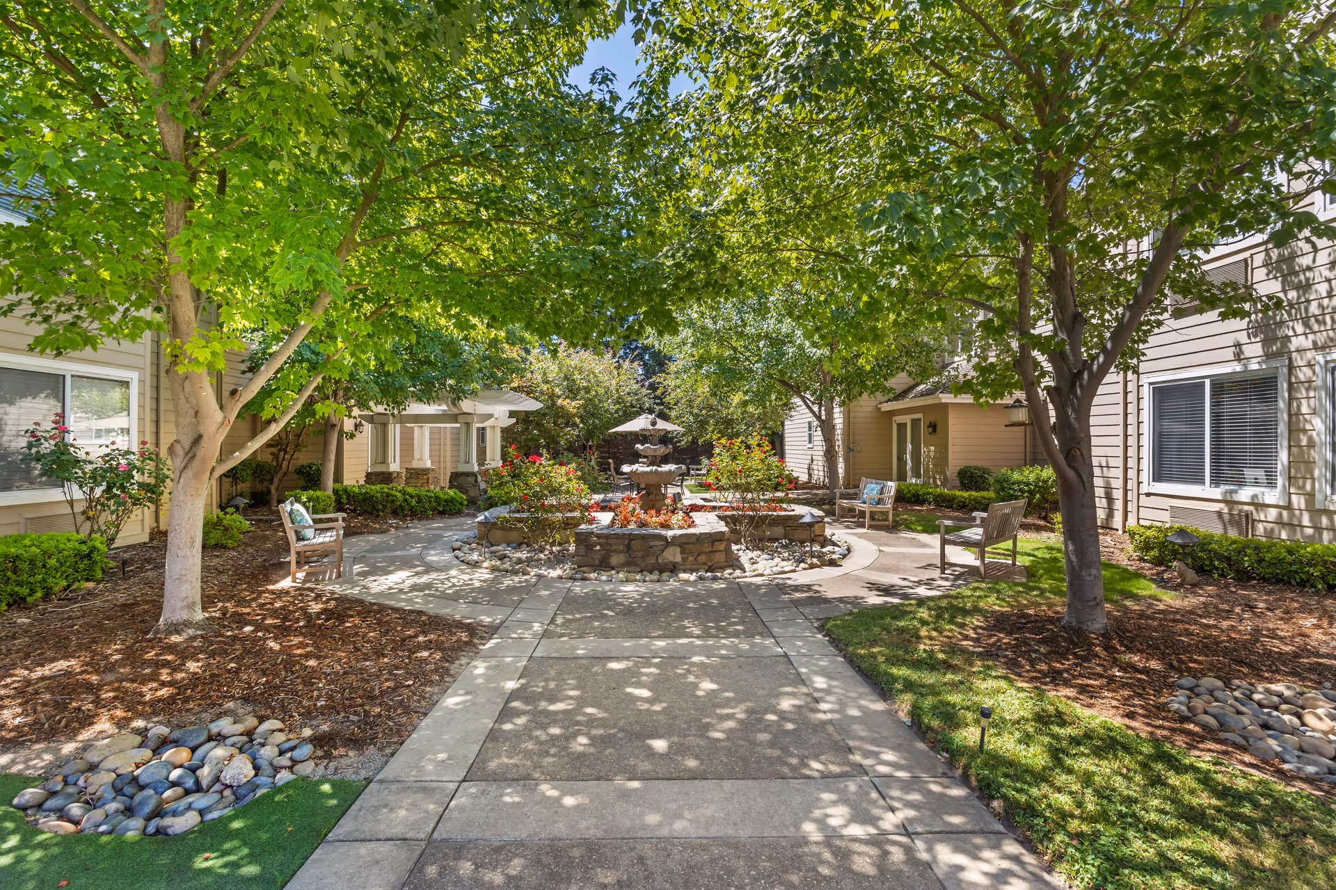 A peaceful outdoor courtyard at Brookdale Lodi featuring a central stone fountain surrounded by flower beds and greenery. The area is shaded by large leafy trees, with benches placed along the paved pathways. The courtyard is bordered by beige residential buildings with windows and doors visible.