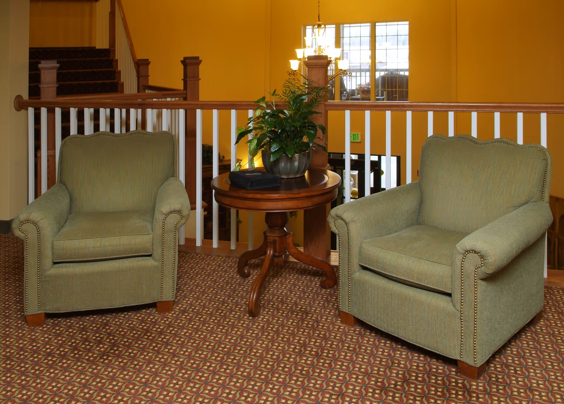 Two green upholstered armchairs with nailhead trim are positioned on a patterned carpeted floor, separated by a round wooden table holding a potted plant and a folded black item. Behind them is a wooden railing with white balusters, and a staircase with a dark carpet runner is visible to the left. A large window and a chandelier are seen in the background.
