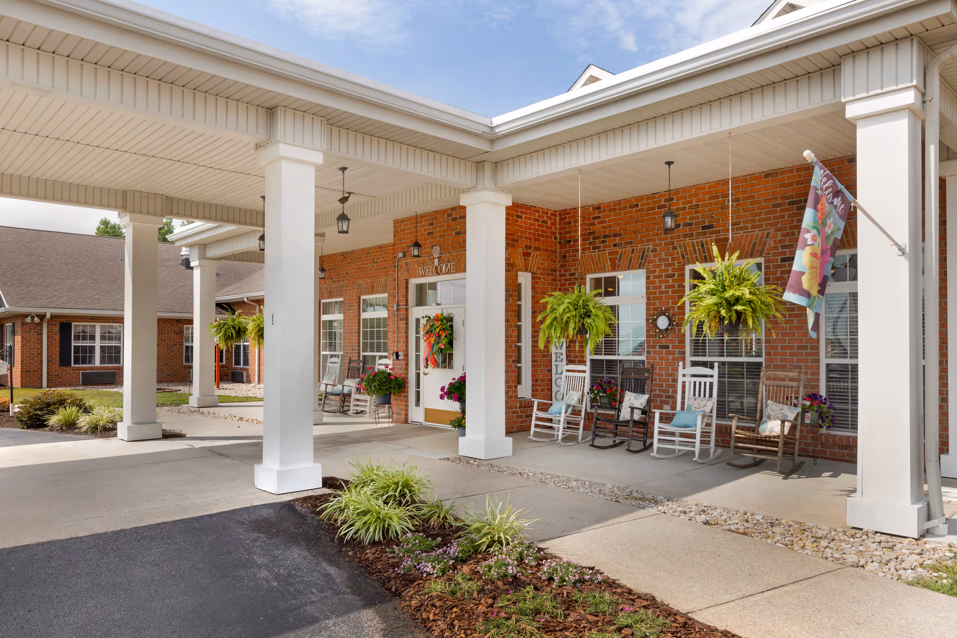 Brick senior living facility entrance under a covered walkway with rocking chairs and plants.