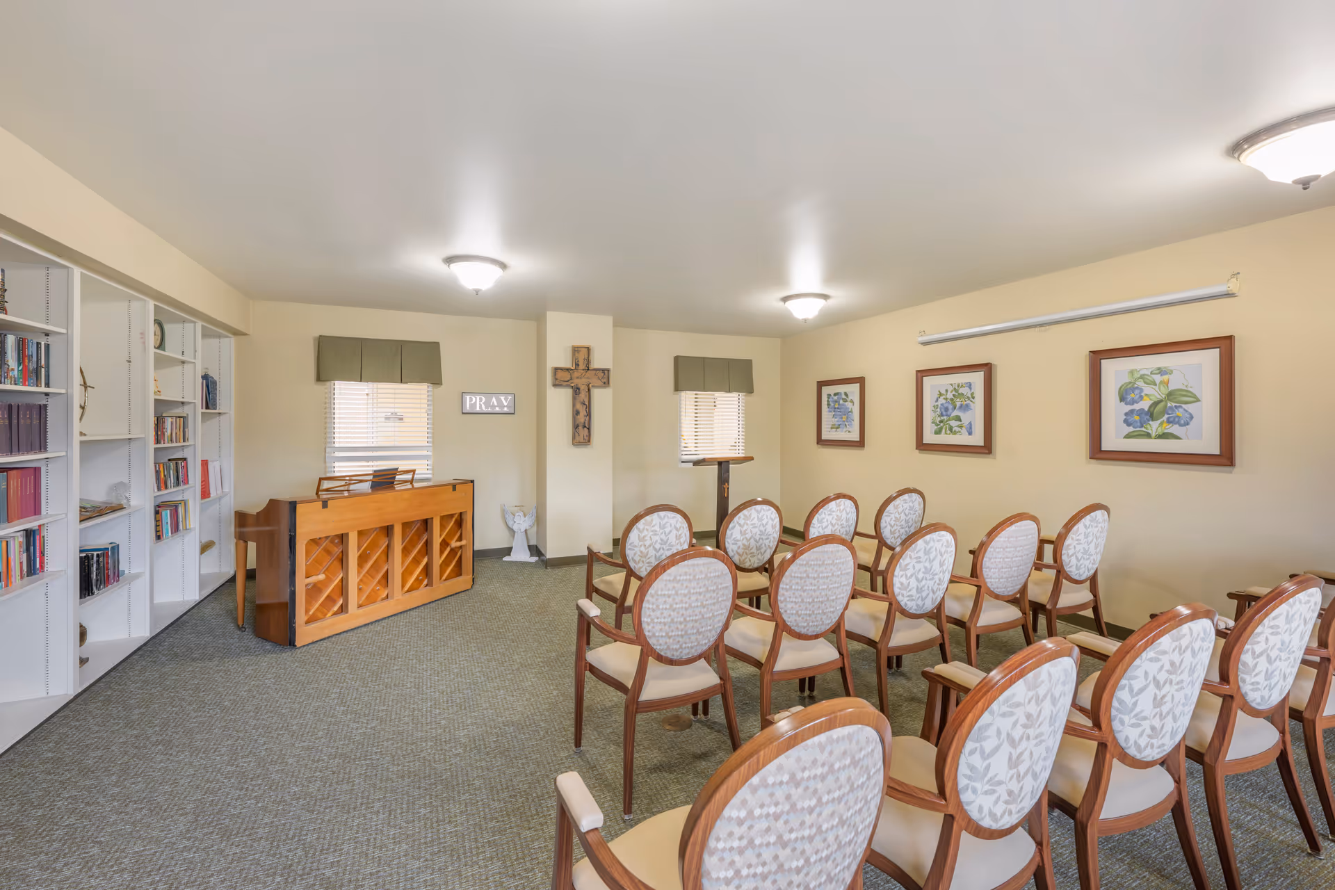 A small chapel or prayer room with rows of wooden chairs upholstered in light patterned fabric facing a wooden piano and a lectern. The walls are light yellow with framed floral artwork and a wooden cross mounted on a central pillar. Two windows with green valances let in natural light, and a white bookshelf filled with books is visible on the left side.