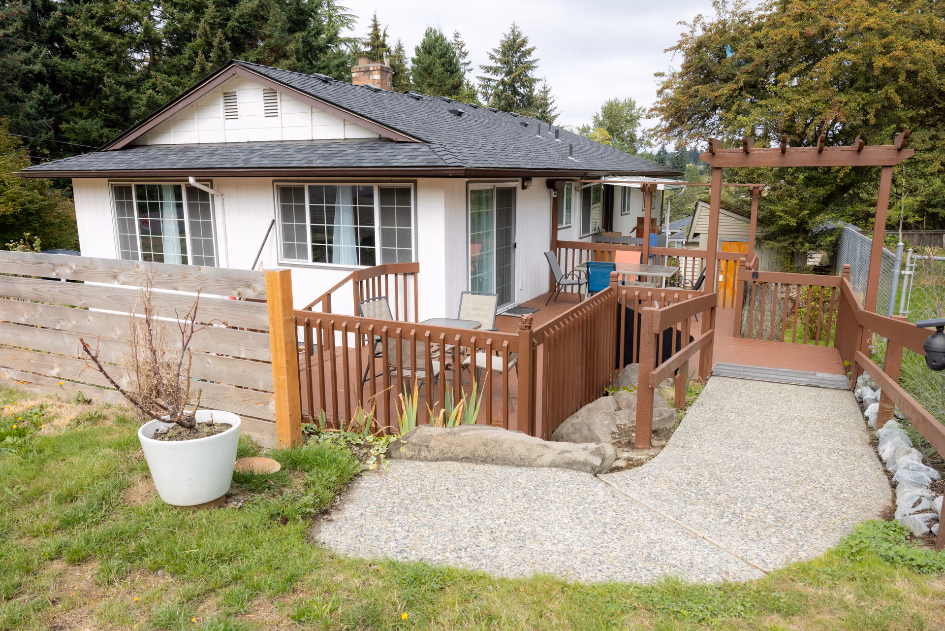 Single-story white house with a wooden deck and ramp leading to a patio area surrounded by grass and trees.