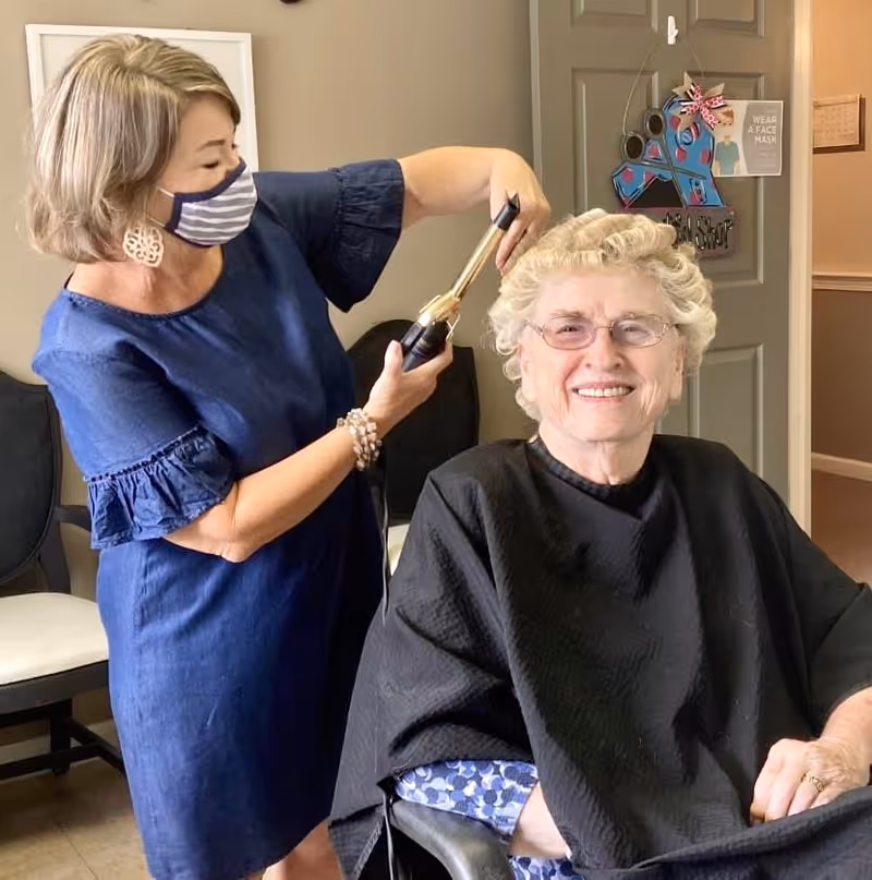 A woman wearing a striped face mask and blue dress is curling the hair of an elderly woman who is seated and smiling, wearing glasses and a black salon cape. They are in a room with chairs and a door decorated with a colorful ribbon and a sign that says 'Wear a Face Mask'.