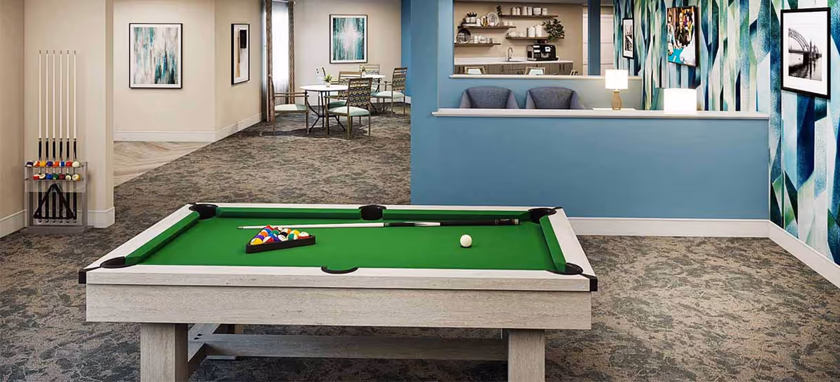 Bright common area featuring a pool table in the foreground with seating, a kitchenette and dining area visible in the background.