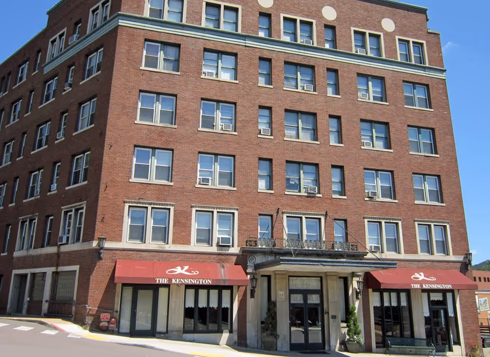 Exterior view of a multi-story brick building with many windows and two red awnings labeled 'The Kensington' above the entrances, under a clear blue sky.
