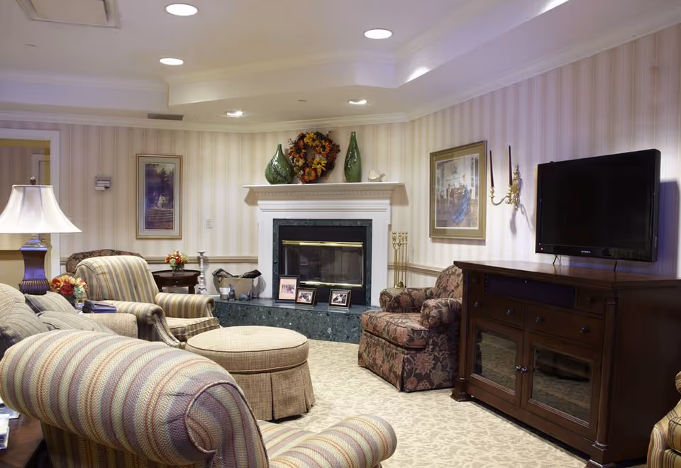Cozy living room with upholstered chairs and ottoman, a fireplace mantel with decorations, and a TV on a wooden cabinet.