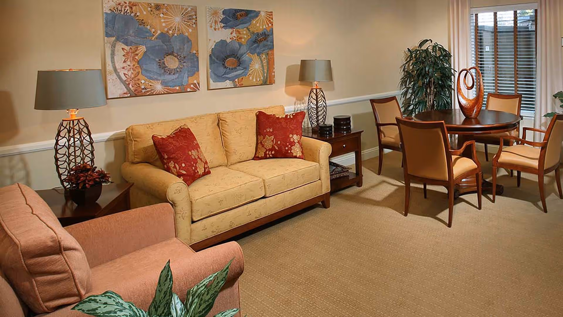 A cozy living room area featuring a beige sofa with two red floral cushions, a pink armchair, two side tables with decorative lamps, a round wooden dining table with four chairs, a large potted plant, and two floral paintings on the wall.