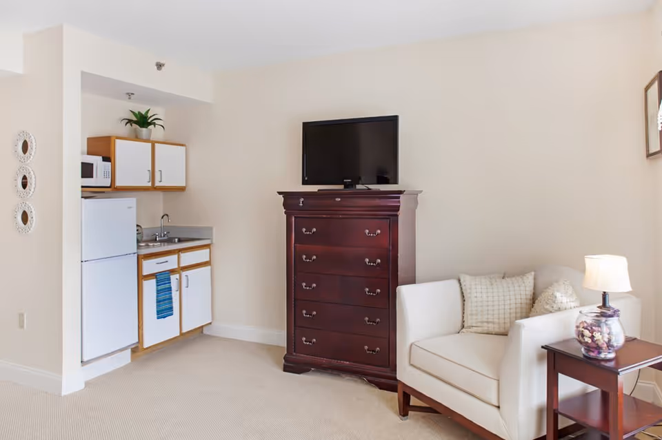 A small living area with a white armchair and two cushions next to a wooden side table with a lamp. A dark wooden chest of drawers with a flat-screen TV on top is against the wall. To the left, there is a compact kitchenette with a white refrigerator, microwave, sink, and cabinets. The walls are light-colored and the floor is carpeted.
