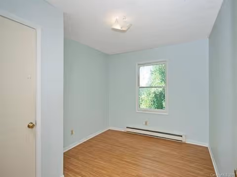 Empty light-blue bedroom with window, baseboard heater, and wood-like floor.