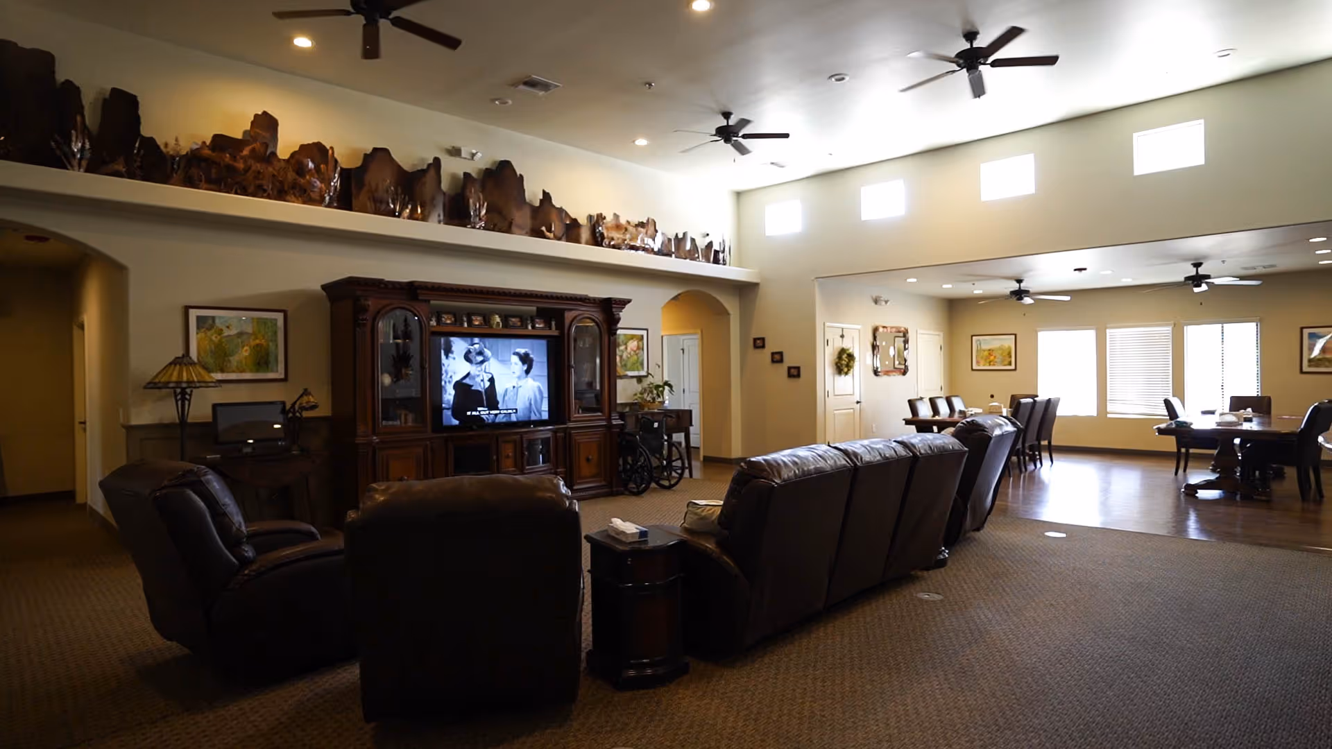 Spacious senior living common room with leather sofas and recliners facing a large entertainment center, high ceilings with fans, and a dining area in the background.