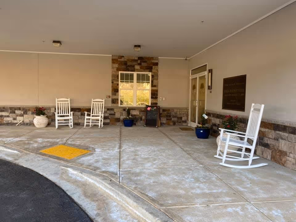 Covered building entrance with white rocking chairs, potted plants, stone accents, and double glass doors.