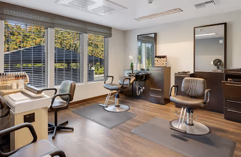A bright and modern salon room with three salon chairs, two large mirrors, and a window with blinds showing greenery outside. The room has wooden flooring and neutral-colored walls with salon equipment and storage cabinets.