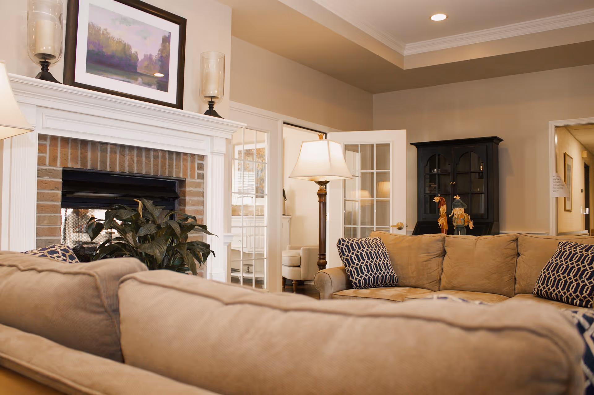 A cozy living room with beige sofas adorned with patterned blue and white pillows, a white fireplace with a brick interior, a framed landscape painting above the mantel, two candle holders on either side of the painting, a floor lamp, a black cabinet with decorative items, and a doorway leading to another room with glass-paneled double doors.