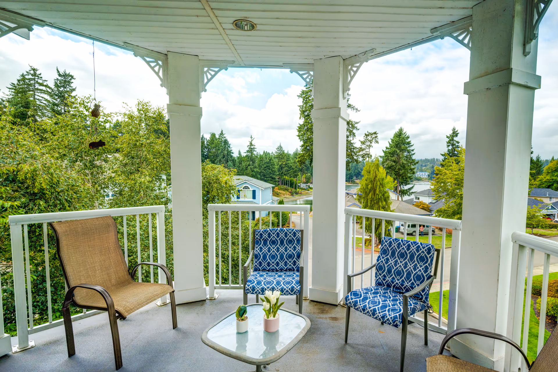Covered outdoor balcony with white railing, featuring two blue patterned chairs, two brown chairs, and a small glass-top table with two small potted plants. The balcony overlooks a neighborhood with trees, houses, and a partly cloudy sky.