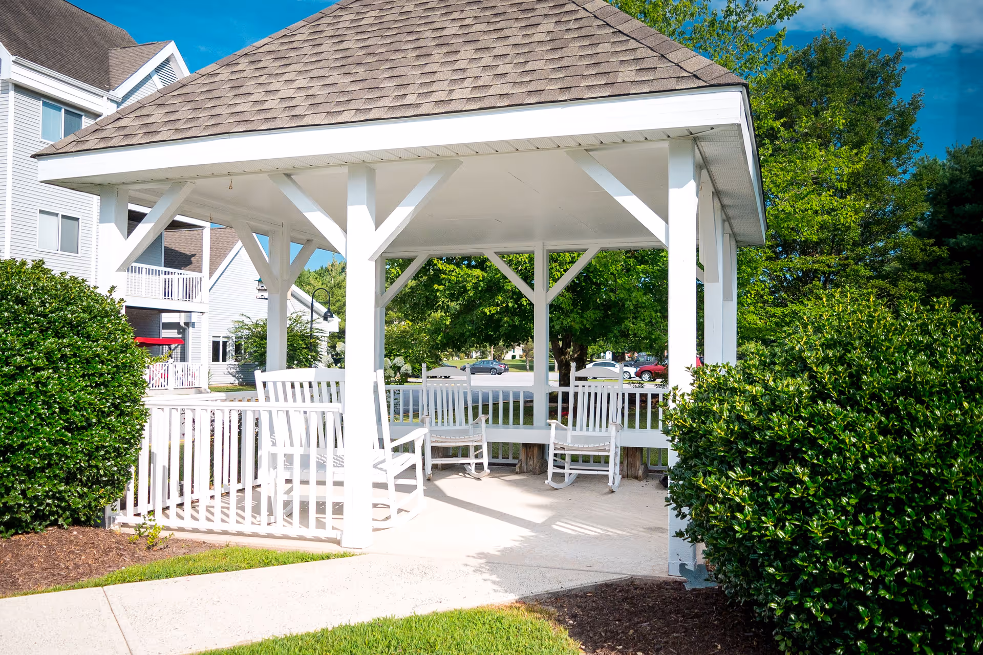 A white wooden gazebo with a shingled roof situated outdoors, surrounded by green bushes and trees. Inside the gazebo are white rocking chairs and a bench. In the background, there are residential buildings and parked cars under a clear blue sky.
