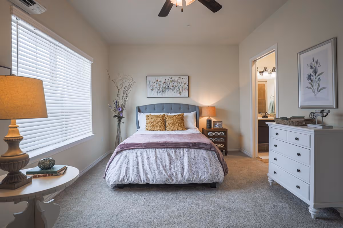 A cozy bedroom with a neatly made bed featuring white and purple bedding and two mustard-colored pillows. There is a nightstand with a lamp and a framed photo to the right of the bed. A large window with white blinds is on the left side, and a white dresser with decorative items is on the right. A doorway leads to a bathroom with a sink and mirror visible. The walls are light-colored, and the floor is carpeted.