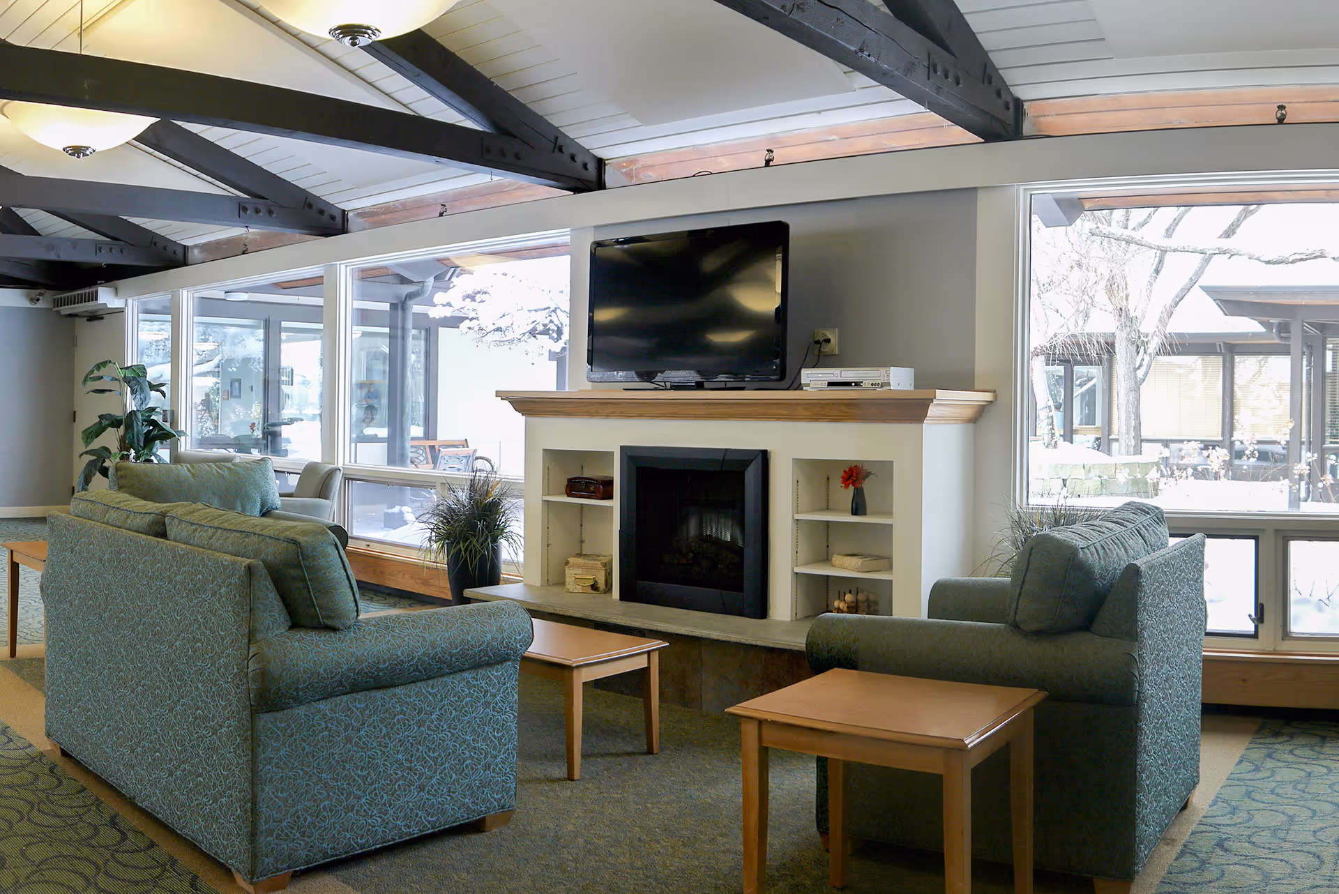 A cozy living room area with two green upholstered sofas facing each other, a wooden coffee table and side table, a fireplace with built-in shelves, and a flat-screen TV mounted above the fireplace. Large windows behind the sofas provide a view of an outdoor area with trees and snow.