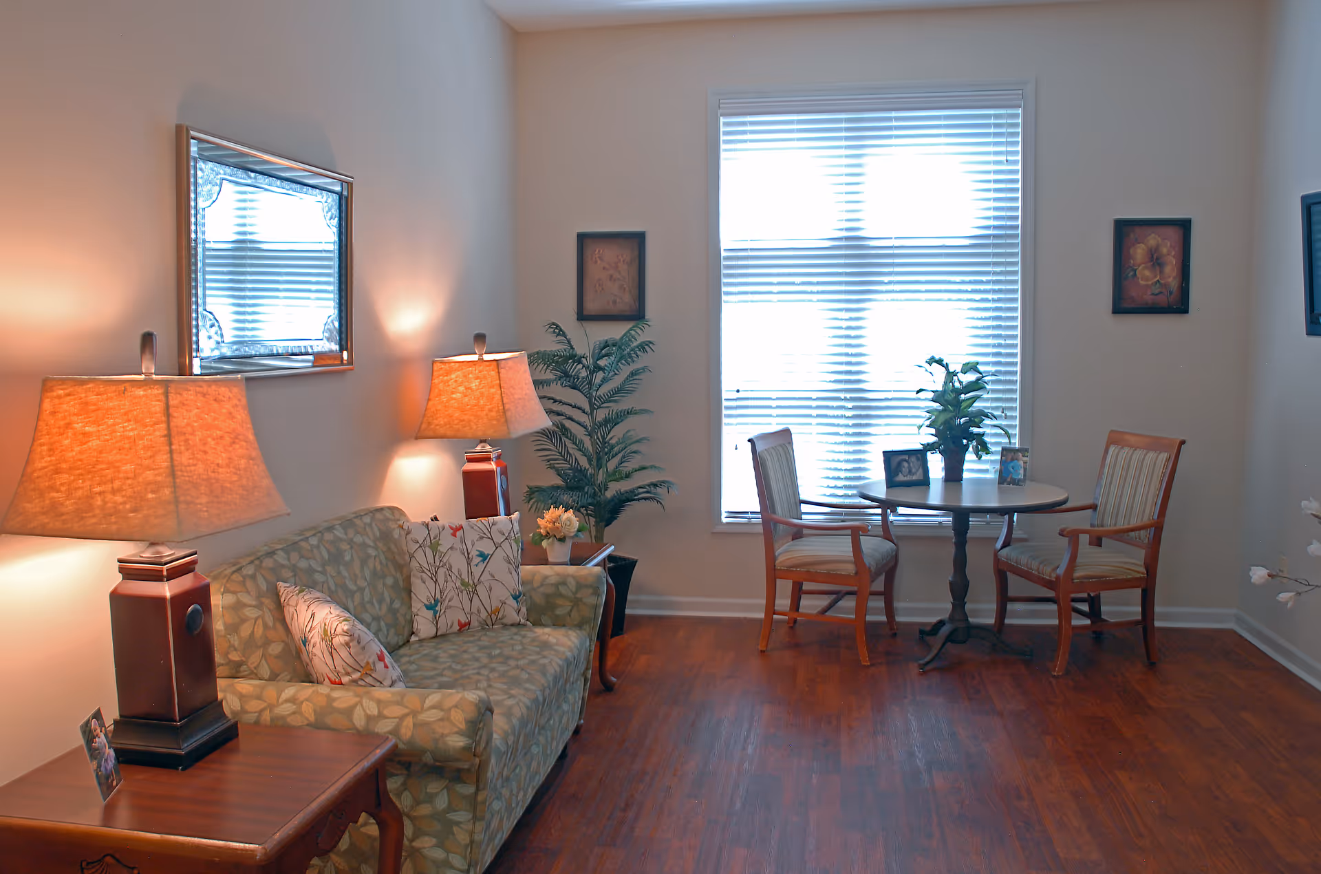 A cozy living room area with a patterned sofa adorned with two decorative pillows, two table lamps with warm light on either side, a wooden side table with a framed photo, a large mirror on the wall, a potted plant in the corner, and a small round table with two wooden chairs near a window with blinds.