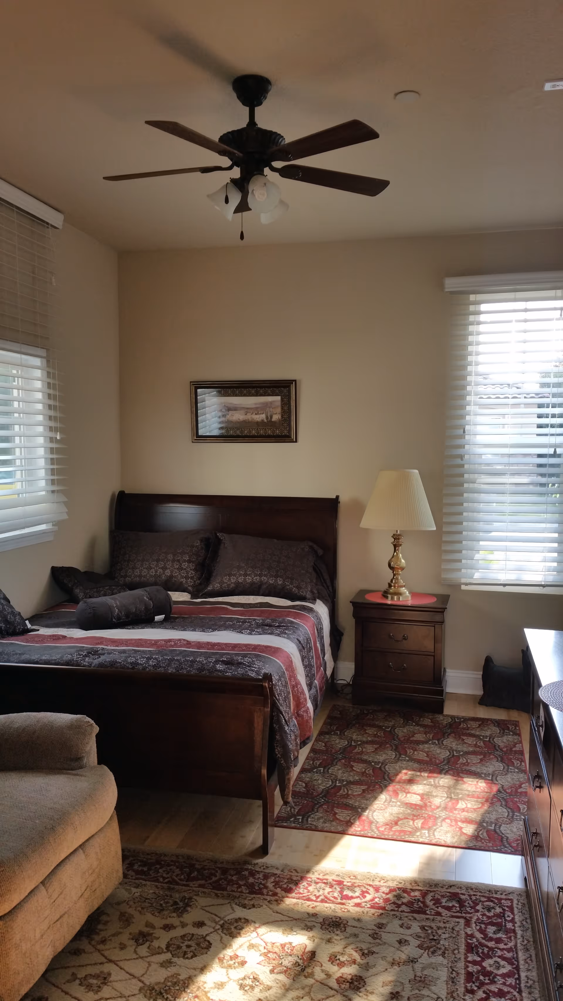 A cozy bedroom with a wooden bed frame, patterned bedding, and several pillows. There is a wooden nightstand with a lamp on it next to the bed. The room has two windows with white blinds allowing natural light to enter. A ceiling fan with lights is mounted on the ceiling. There are patterned rugs on the wooden floor and a beige armchair partially visible in the foreground.