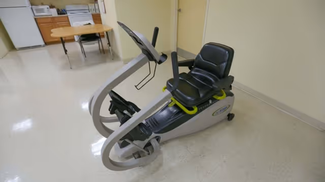 A recumbent exercise bike with a black seat and gray frame is placed in a room with a tiled floor and light-colored walls. In the background, there is a small wooden table with chairs, a white refrigerator, and a microwave on a countertop.