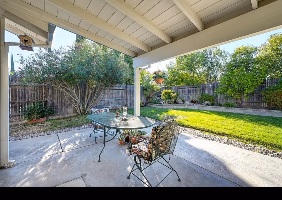 Covered backyard patio with a glass-top table and two metal chairs overlooking a lawn and fenced garden.