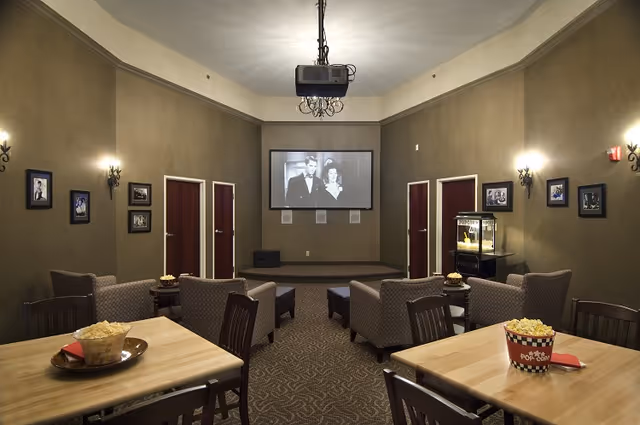 A cozy community theater-style lounge with a projector screen at the front, upholstered chairs and tables topped with popcorn, and a popcorn machine.