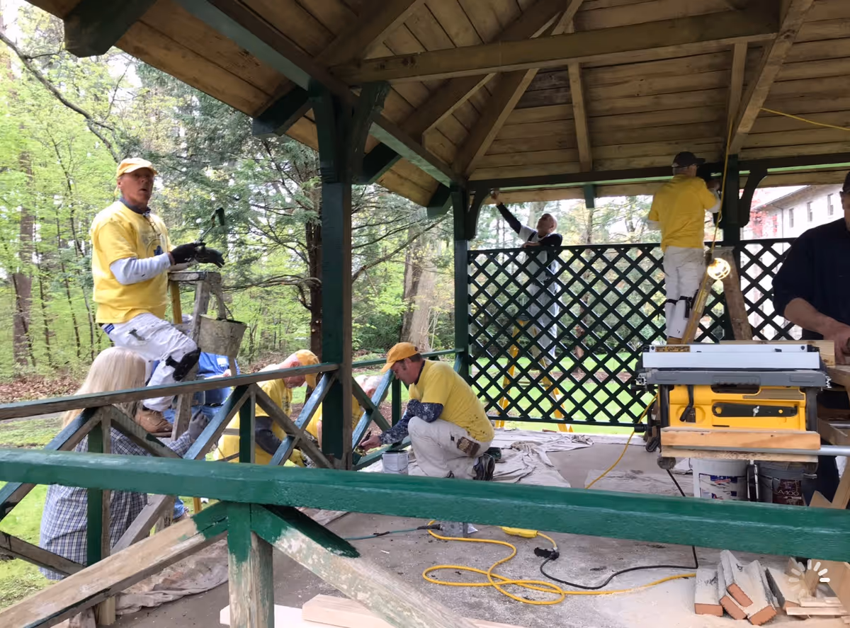 Several people wearing yellow shirts and white pants are working together to paint and renovate a wooden gazebo with a green railing and lattice panels, surrounded by trees and greenery.