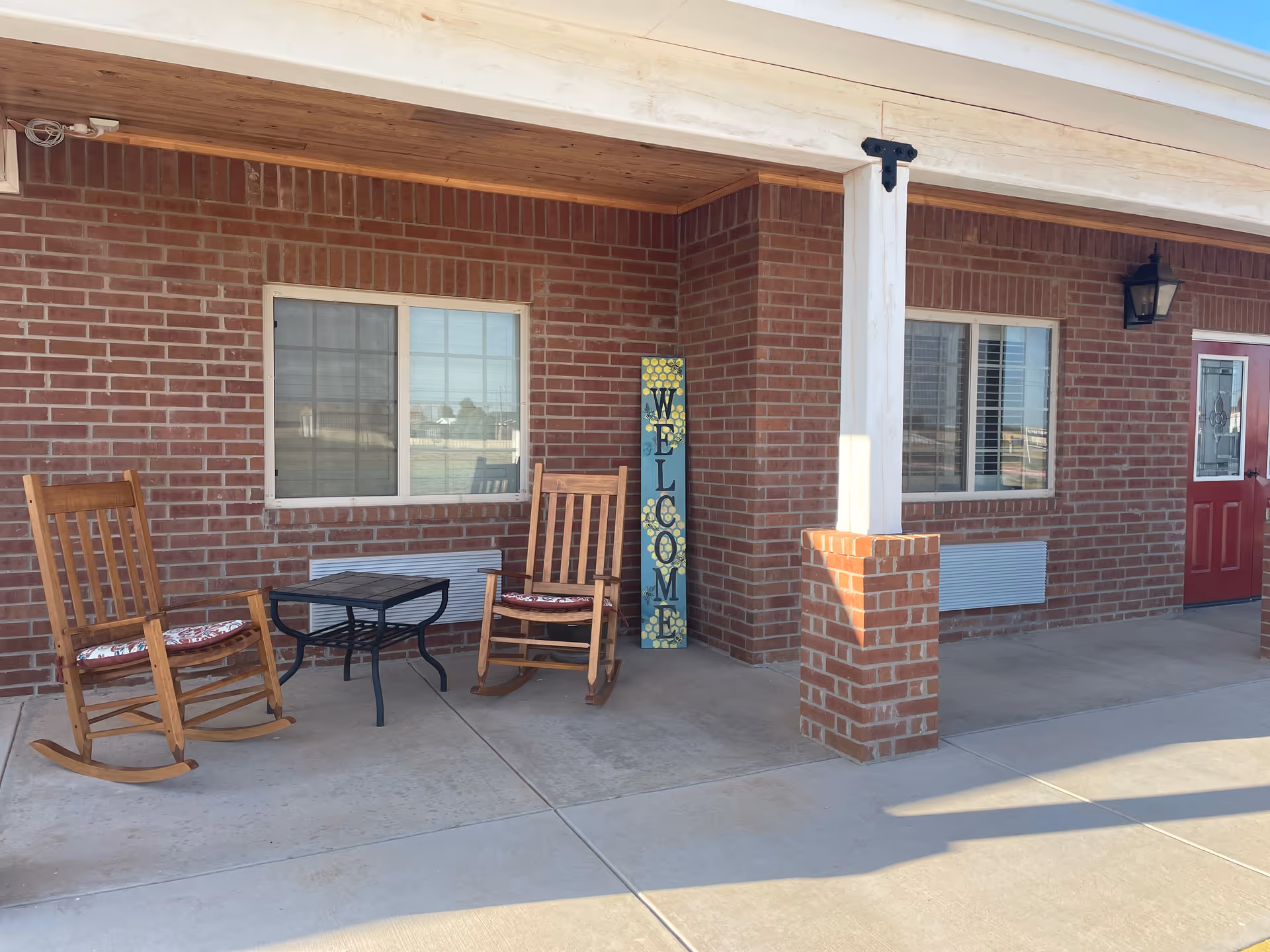 Covered brick front porch with two wooden rocking chairs, a small table, and a vertical "WELCOME" sign next to a red entry door.