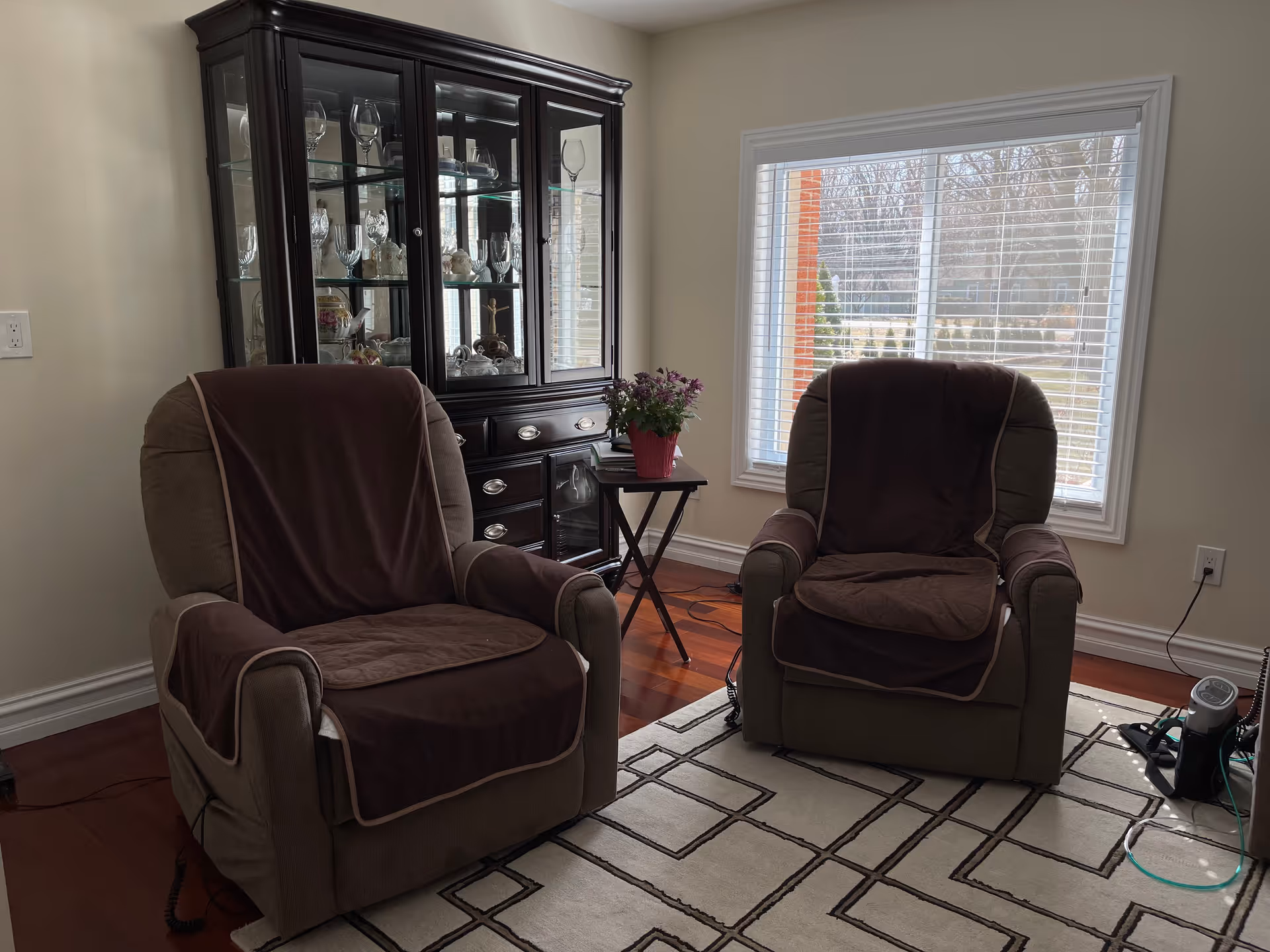 A cozy living room area with two cushioned armchairs covered with brown protective covers, a small side table with a potted plant, a dark wooden glass-front cabinet displaying glassware and decorative items, a large window with white blinds letting in natural light, and a patterned area rug on a wooden floor.