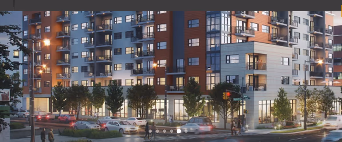 Exterior view of a modern multi-story residential building at dusk with lit windows, balconies, trees along the sidewalk, cars on the street, and pedestrians crossing at an intersection.