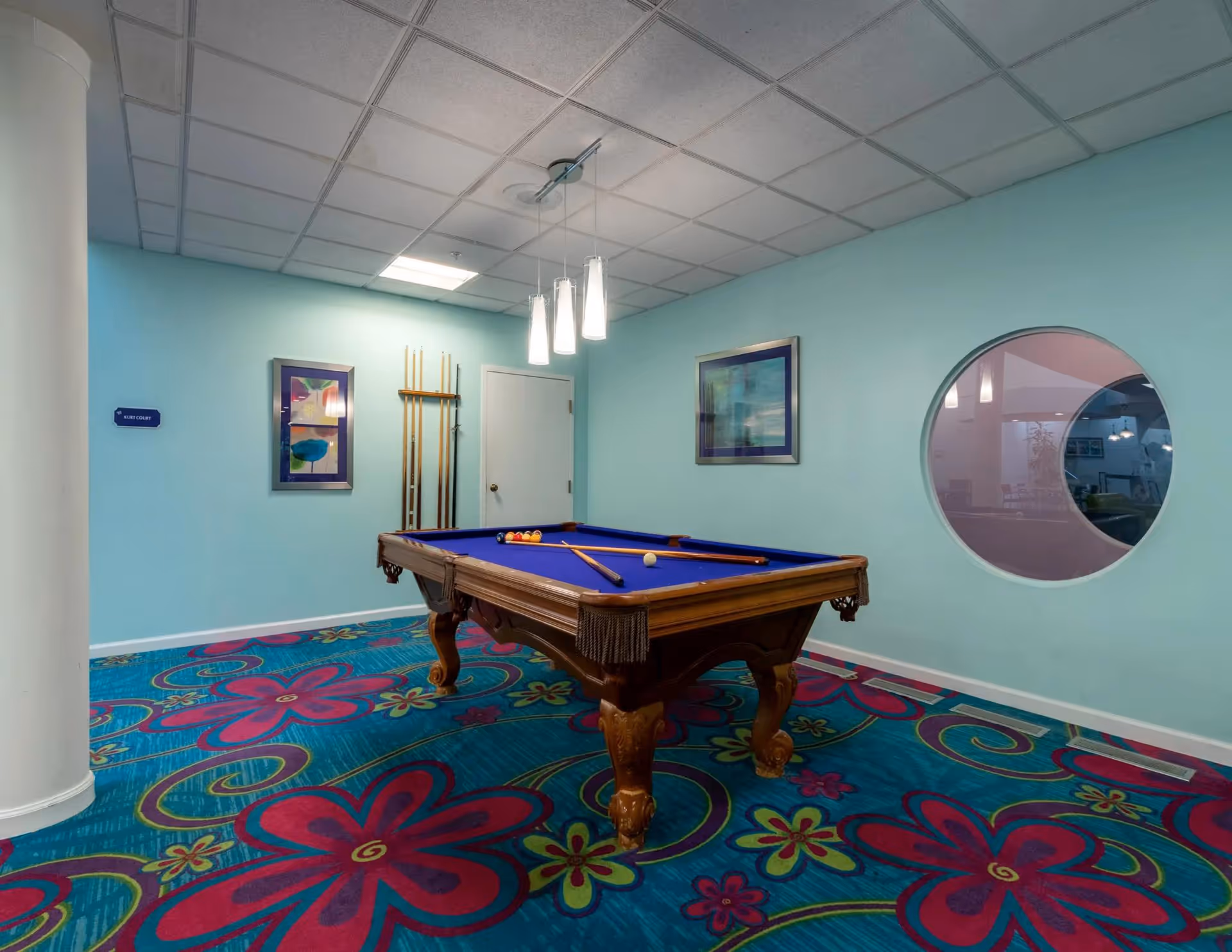 A recreational room with a pool table featuring a blue felt surface. The room has light blue walls, colorful carpet with large floral patterns in red, pink, and green, and a ceiling with white tiles and hanging pendant lights. There are two framed pictures on the walls and a circular window showing a glimpse of an adjacent room.
