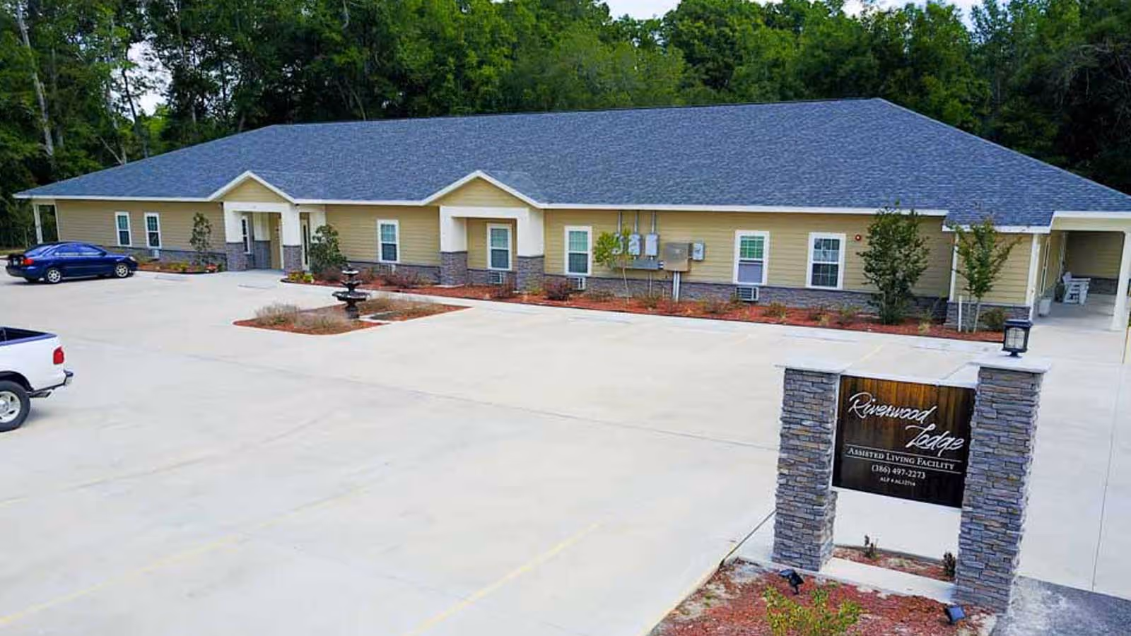 Exterior view of a single-story assisted living facility building with beige siding and a dark gray roof, surrounded by trees. There is a parking lot in front with a few parked cars and a stone sign that reads 'Riverwood Lodge Assisted Living Facility'.