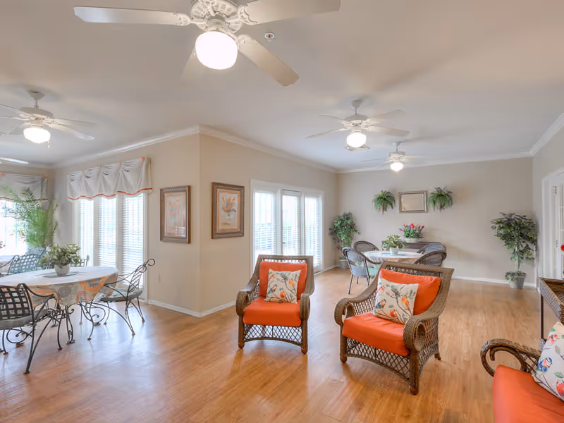 A bright and spacious common area with wooden flooring, featuring wicker chairs with orange cushions and floral pillows, round tables with metal chairs, ceiling fans with lights, potted plants, and framed artwork on beige walls.
