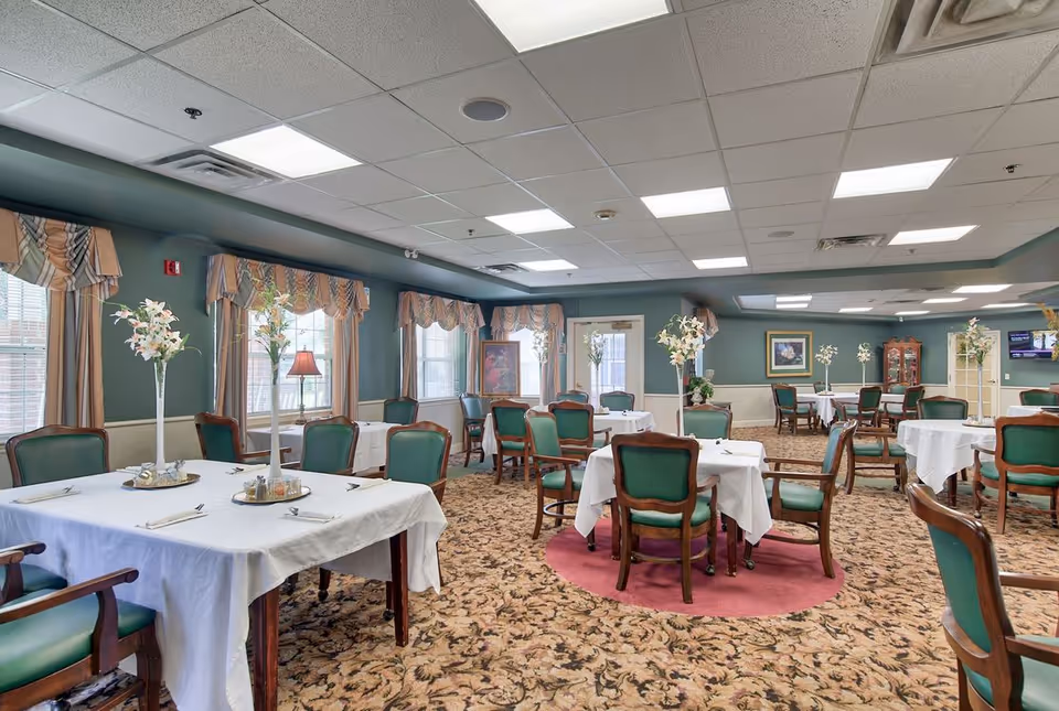 A spacious dining room with multiple tables covered in white tablecloths, each set with green cushioned wooden chairs and floral centerpieces. The room has patterned carpet, green walls with beige wainscoting, large windows with valances, and ceiling lights providing bright illumination.