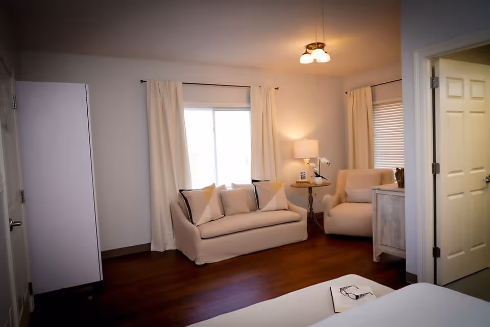 A cozy living room area with a beige loveseat and armchair, both adorned with light-colored pillows. A small round wooden side table with a lamp, books, and a small plant sits between the two seating pieces. The room has hardwood flooring, white walls, and two windows with white curtains. A door is open to an adjacent room, and part of a bed with a book and glasses on it is visible in the foreground.
