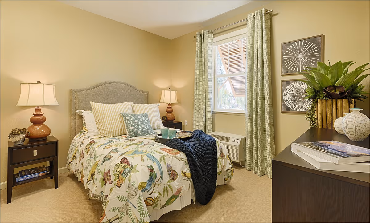 A cozy bedroom with a bed featuring a colorful bird and butterfly patterned bedspread, multiple pillows, and a dark blue knitted throw. There are two bedside tables with matching lamps, a window with light green curtains, and a dark dresser with decorative plants and books. The walls are painted light beige.