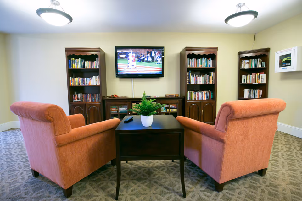 A cozy living room area with two orange upholstered armchairs facing a dark wooden coffee table with a small potted plant. Behind the table, there are three wooden bookshelves filled with books and a flat-screen TV mounted on the wall showing a baseball game. The room has patterned carpet flooring and two ceiling lights.