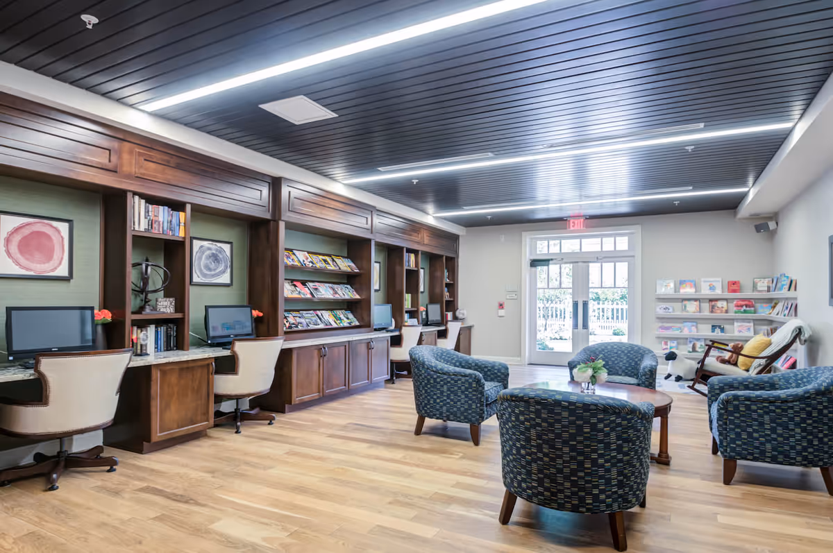 A bright and spacious common area in Arbor Terrace Morris Plains featuring a row of wooden desks with computers and chairs along one wall, a magazine rack, and comfortable blue patterned armchairs arranged around a wooden coffee table. The room has a modern dark ceiling with linear lighting, light wood flooring, and a glass door leading outside. There is also a small bookshelf with children's books and a rocking chair with a teddy bear in the corner.