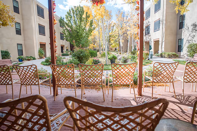 View of an outdoor courtyard area at Brookdale Santa Fe with several empty metal lattice chairs arranged on a brick patio facing a garden with trees and shrubs. The courtyard is surrounded by multi-story beige buildings with windows, and the trees have autumn-colored leaves.