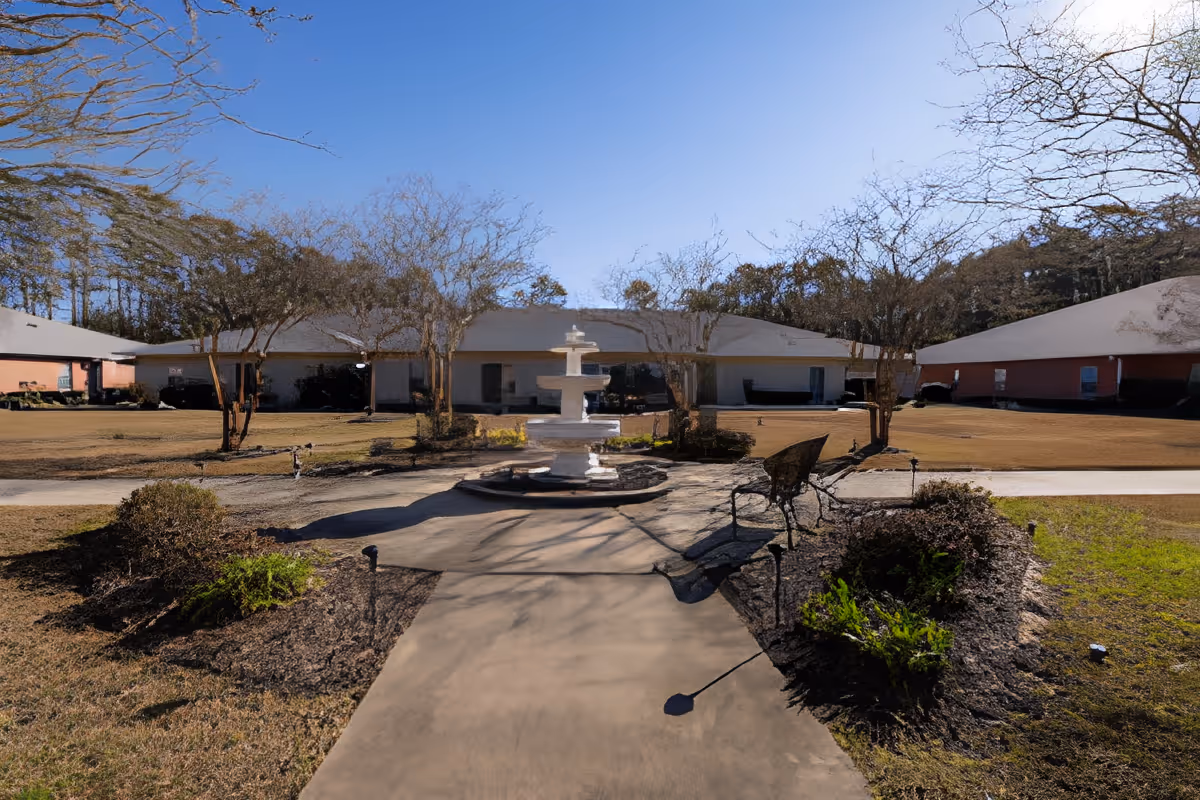 Outdoor courtyard area with a concrete pathway leading to a white multi-tiered fountain in the center. The courtyard is surrounded by low bushes and small trees with leafless branches. There are benches on either side of the pathway. Single-story buildings with light-colored walls and gray roofs are visible in the background under a clear blue sky.
