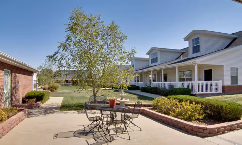 Outdoor patio area at Teal Lake Senior Living with a metal table and chairs set on a concrete surface, surrounded by landscaped bushes and a tree. In the background, there are white residential buildings with porches and a clear blue sky.