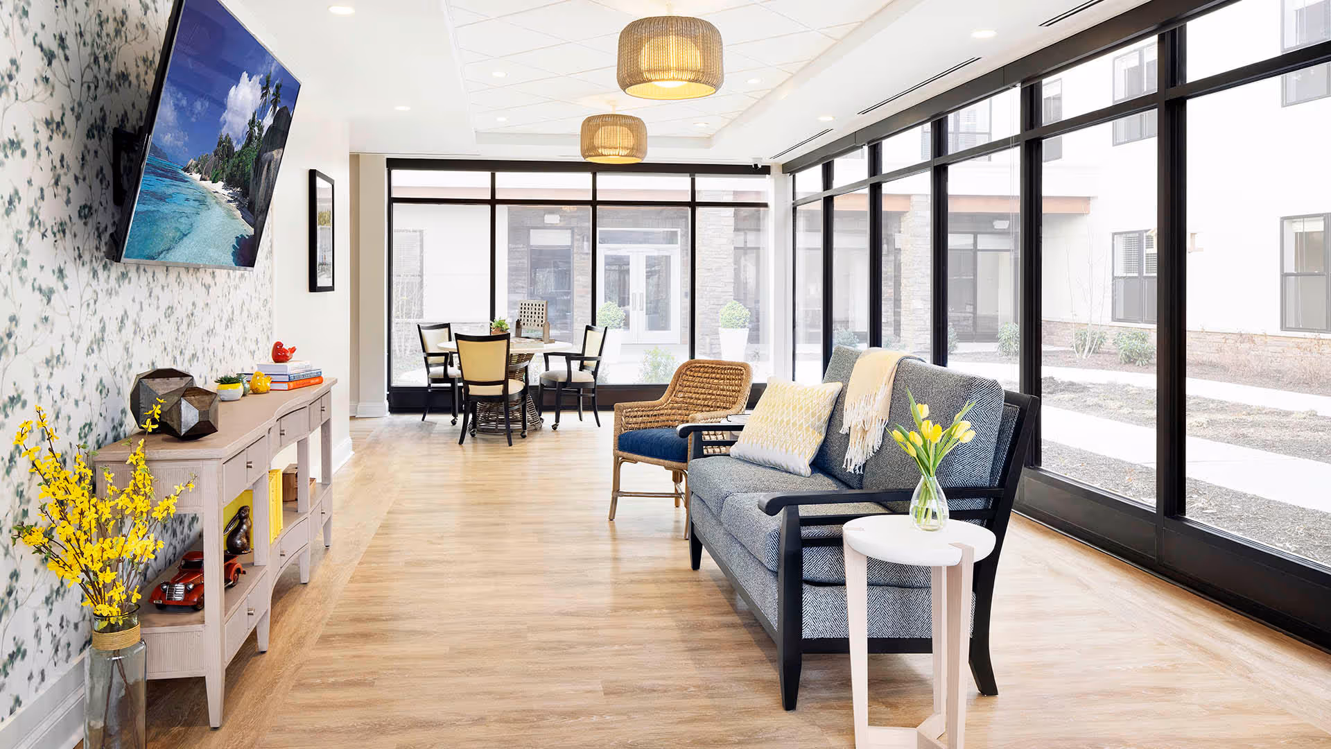 A bright and modern living area with large floor-to-ceiling windows letting in natural light. The room features a gray sofa with cushions and a throw, a small white side table with a vase of yellow tulips, a wicker chair, and a dining table with chairs in the background. A flat-screen TV is mounted on a floral-patterned wall above a console table decorated with books, plants, and decorative items. The floor is light wood, and two pendant lights hang from the ceiling.