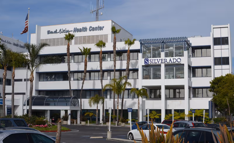 Front exterior of a multi-story white building labeled 'Silverado' and 'Beach Cities - Health Center' with palm trees and parked cars in front.